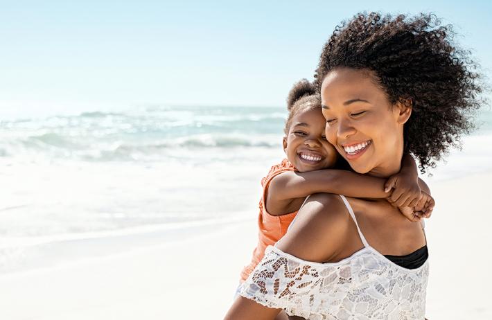 A mom giving her daughter a piggyback ride on the beach, both smiling